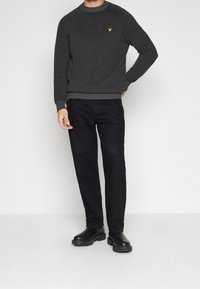 Dark grey knitted sweater with ribbed collar and cuffs, featuring a small yellow logo. Paired with black straight-leg trousers and black boots.