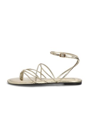 Sandalia plana de oro metálico con finas tiras entrelazadas, lazo para el dedo y correa en el tobillo con hebilla pequeña sobre una suela negra.