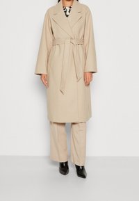 Abrigo beige de mezcla de lana con cuello ancho, cintura ceñida con cinturón y mangas largas y sueltas. Llevado sobre pantalones beige y botas negras puntiagudas.