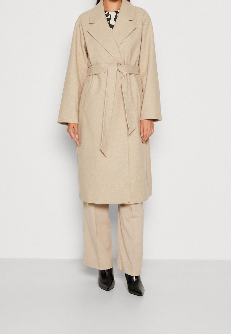 Abrigo beige de mezcla de lana con cuello ancho, cintura ceñida con cinturón y mangas largas y sueltas. Llevado sobre pantalones beige y botas negras puntiagudas.