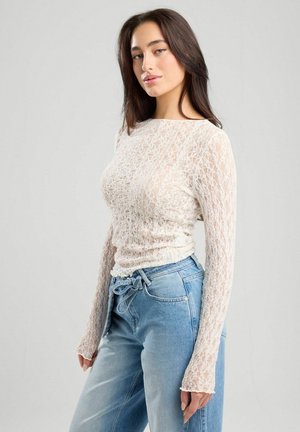 Lichte, doorschijnende witte kanten top met lange mouwen en een textuurpatroon, gecombineerd met hoogsluitende blauwe spijkerbroek met een trekkoord in de taille.