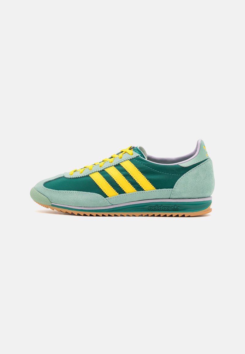 Låga Adidas-skor i teal och gult med mockaöverdrag, gula snörning, signaturens tre ränder och gummi sula på vit bakgrund.