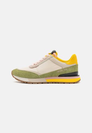 Sneaker met zacht beige leer, groene suède accenten, roze veters, zwarte en gele zool, met een strak, modern ontwerp en contrasterende texturen.