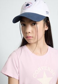 Hvid og navy baseballcap med et stjerne-logo, båret med en lyserød T-shirt med et stort stjerne grafik. Glat stofstruktur.