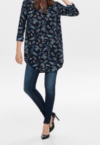 Mörkblå tunikaöverdel med paisleymönster, med button-down krage, trekvartsärmar och krökt fåll, som är kombinerad med mörkblå jeans.