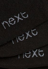 Tissu brun foncé avec trois bandes superposées portant le mot "next" en couture blanche. La texture est tissée avec des détails fins.