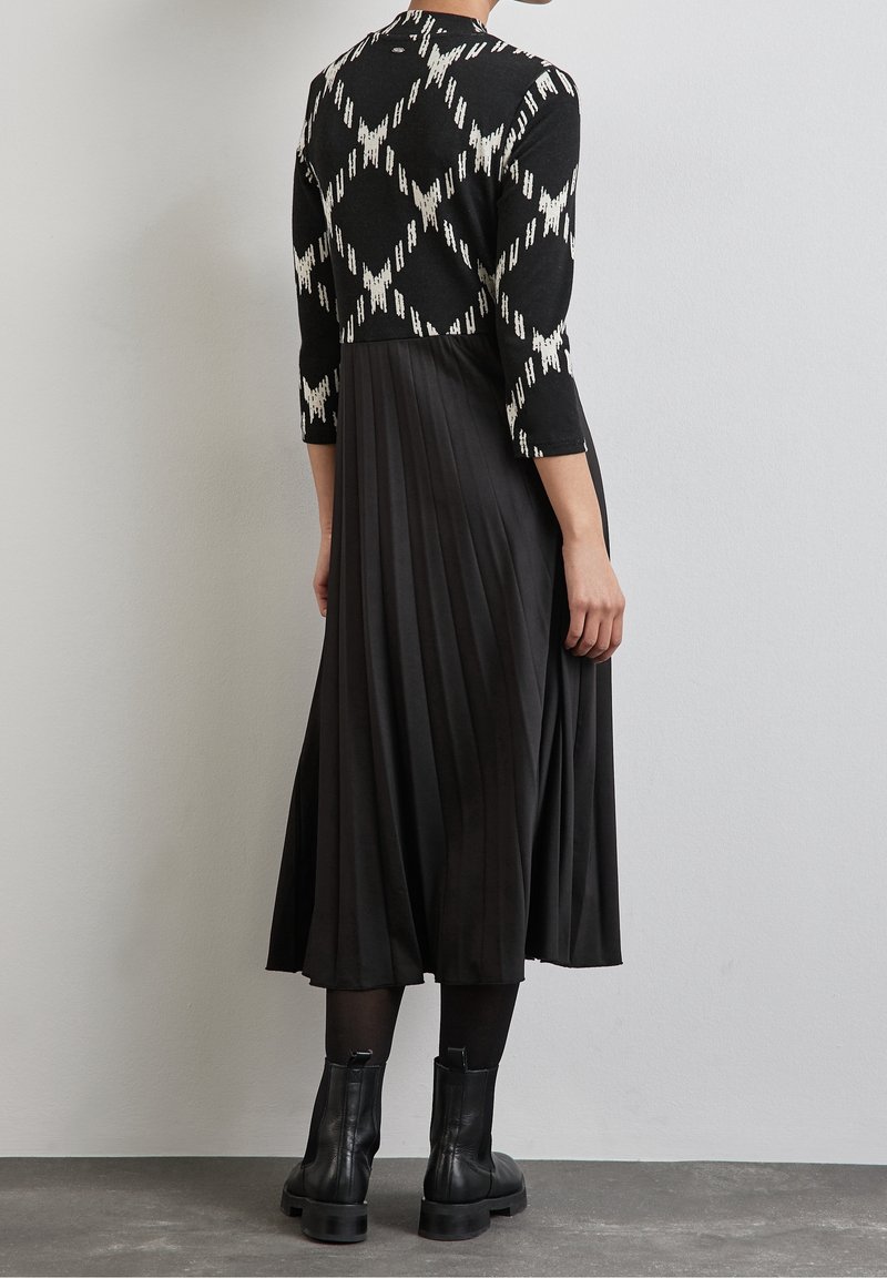 Femme portant un haut à motif noir et blanc avec une jupe midi noire plissée, des collants noirs et des bottines noires, debout contre un mur uni.
