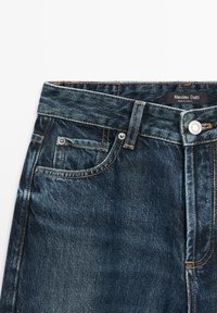 Dark blue denim jeans with a mid-rise waist, highlighted stitching, five pockets, and a metal button closure. Brand label visible.