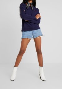 Tmavě modrá mikina s logem, džínové šortky s ohrnutými okraji a bílé kotníkové boty, stylizované s černobílým šátkem kolem krku.