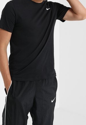 Homme portant un t-shirt Nike noir et un pantalon de survêtement Nike noir, debout avec une main dans la poche et l'autre levée près de la tête.
