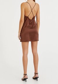 Robe mini en satin marron avec un dos nu et des bretelles croisées. Caractérisée par une texture lisse et une silhouette ajustée, portée avec des sandales noires à lanières.