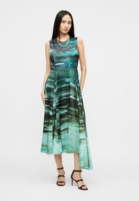 Γυναίκα με μακριά σκούρα μαλλιά που φοράει αμάνικο μακρύ φόρεμα σε απόχρωση τιρκουάζ με μαύρα σχέδια και μαύρες ψηλοτάκουνες σανδάλες, στέκεται μπροστά σε λευκό φόντο.