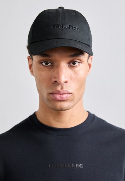 Sort baseball cap med broderet logo, båret med en sort t-shirt med korte ærmer, der viser et subtilt præget mærkenavn. Glat stofoverflade.