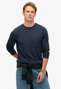 Camiseta de manga larga azul marino en tejido suave, de corte relajado. Presenta mangas enrolladas y un suéter de cuadros verdes casual atado en la cintura.