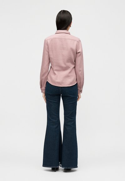 Γυναίκα με μακριά σκούρα μαλλιά που φορά ανοιχτό ροζ μακρυμάνικη μπλούζα και σκούρα μπλε καμπάνα τζιν, στέκεται πλάτη σε λευκό φόντο.