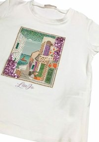 T-shirt bianco in cotone con una grafica decorativa impreziosita da paillettes colorate, che rappresenta una scena costiera con edifici e una barca, firmata LiuJo.