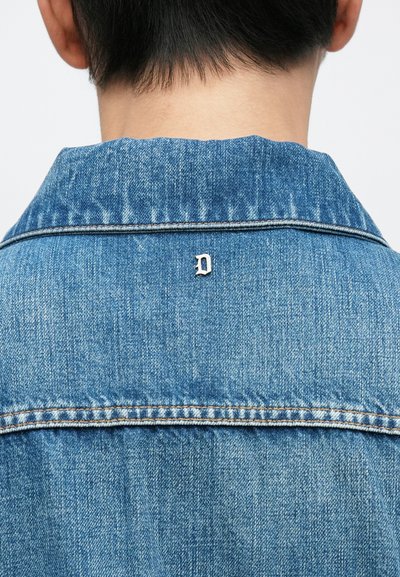 Veste en denim bleu clair avec un col classique. Équipée de ferrures argentées et d'un petit accent métallique en forme de "D" près de l'encolure.