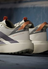 Ζευγάρι μπεζ και γκρι παπούτσια τρεξίματος Adidas Adizero Evo SL ATR με πορτοκαλί λουριά έλξης πάνω σε υφή μπλε επιφάνειας.