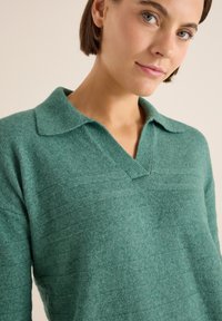 Grüner Pullover aus strukturiertem Stoff, mit einem Kragen-Ausschnitt und horizontalen Streifen über der Brust. Weiches, leichtes Material.