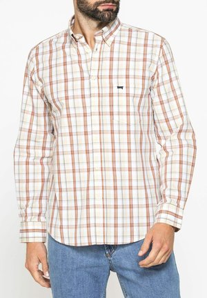 Uomo con una camicia a quadri beige, arancione e bianca a maniche lunghe abbottonata e jeans blu, in piedi su uno sfondo bianco.