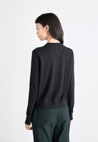Sweatshirt noir à manches longues, avec des poignets et un ourlet côtelés, détail de décolleté en V au dos, associé à un pantalon vert.