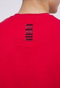T-shirt di cotone rossa con scollatura rotonda. Presenta sette righe orizzontali nere sulla parte superiore della schiena, creando un dettaglio a contrasto.