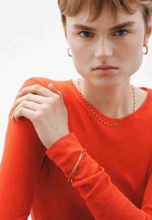 Top naranja de manga larga con mangas ajustadas, collar dorado en capas y una pulsera dorada delgada y retorcida en la muñeca, mostrando texturas suaves.