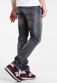 Hombre vestido con jeans negros desgastados y zapatillas maroon de New Balance, de pie con las piernas cruzadas sobre fondo blanco.