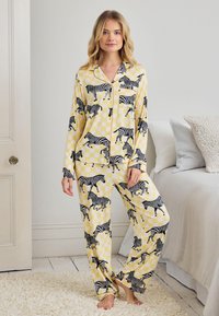 Nainen seisoo paljain jaloin keltaisissa ruudullisissa pyjamassa, joissa on mustavalkoisia seepraprinttejä, pehmeästi valaistussa makuuhuoneympäristössä.