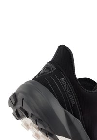 Mustekkiä urheilujalkine musta, jossa on teksturoitu päällinen, logo ja uurteiset yksityiskohdat. Ulkopohjassa on uramalli, joka tarjoaa pidon.