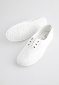 Chaussures en toile blanche à motif en dentelle avec une semelle en caoutchouc, comportant cinq œillets et une surface texturée. Design simple et minimaliste.