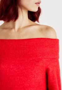 Maglione rosso scollo a spalla con colletto a coste, texture morbida e vestibilità ampia. Presenta un colore vivido e uniforme senza motivi.