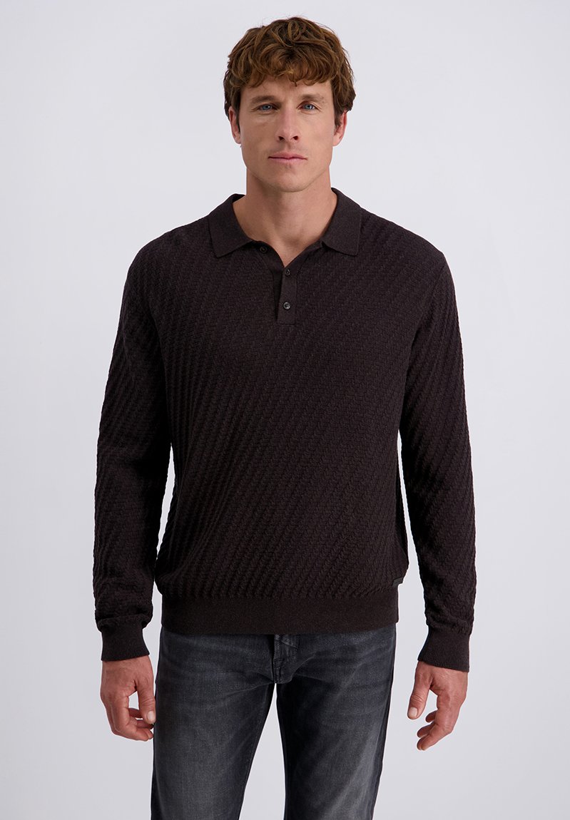 Mann in einem dunkel strukturierten langärmligen Poloshirt und ausgebleichten schwarzen Jeans, der vor einem schlichten hellen Hintergrund steht.