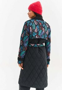 Prešívaný kabát s čiernym základom a farebnými kvetinovými a listovými vzormi na hornej časti, doplnený opaskom na zaväzovanie a manžetami na gombíky.