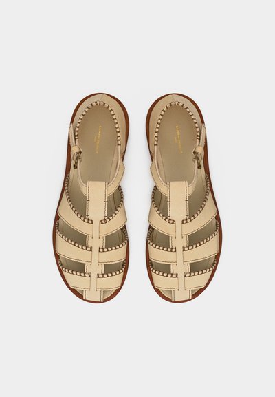 Ζευγάρι μπεζ δερμάτινα πλεκτά σανδάλια με λουράκια στον αστράγαλο και καφέ σόλες, εμφανιζόμενα από πάνω σε απλό φόντο.