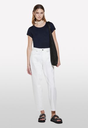 Marineblaues, tailliertes T-Shirt kombiniert mit hochtaillierten, weißen Jeans. Eine schwarze Netzhandtasche und schwarze Sandalen mit Akzenten runden das Outfit ab.