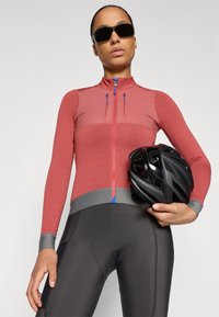 Rød cykeljakke med lynlås foran og ribbet tekstur, parret med sorte cykelbukser og en sort hjelm, holdt i den ene hånd.