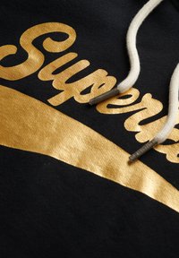 Tejido negro con letras doradas "Superdry" y una franja curva dorada. Presenta cordones crema con puntas metálicas.