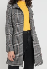 Grå- og sortternet frakke med krave-design, knaplukning og lange ærmer, parret med en gul ribbet top og sorte bukser.
