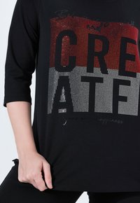 Černé tričko s tříčtvrtečními rukávy, na kterém je velký grafický design z červených a bílých kamínků, který zobrazuje slovo "CREATE" s dalším textem pod ním.
