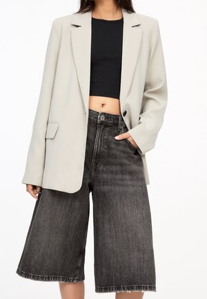 Frau trägt einen beigen Blazer, ein schwarzes Cropped Top und weite schwarze Denim-Culottes mit ausgefranstem Saum, Hand in der Tasche.