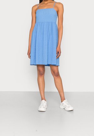 Femme portant une robe d'été texturée bleu clair longueur genou avec fines bretelles et baskets blanches épaisses, debout devant un fond uni.