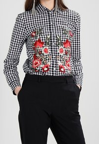 Randig skjorta i svart och vitt med röd blomsterbroderi på bröstet, knapprad, och långa ärmar. Bärs med svarta byxor.