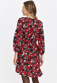 Kobieta widziana z tyłu, ubrana w długą sukienkę z rękawami do kolan w czerwono-białe i zielone kwiatowe wzory oraz czarne rajstopy.