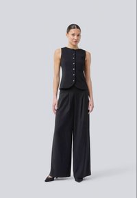 Chaleco negro a medida con cierre de botones combinado con pantalones negros de pierna ancha. Tejido suave, diseño minimalista y silueta elegante.