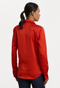 Camisa de seda vermelha de manga longa com gola pontuda, punhos com botão e bainha curva. Textura suave, ajuste feito à medida e design minimalista.