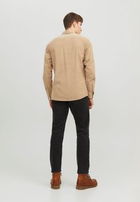 Camisa beige de botonadura con cuello y mangas largas, combinada con pantalones negros ajustados y botas de cuero marrón. Detalles de tela simples y texturizados.
