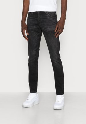 Mann trägt eng anliegende schwarze Jeans, weiße Sneakers und ein weißes Hemd und steht vor einem schlichten hellen Hintergrund.