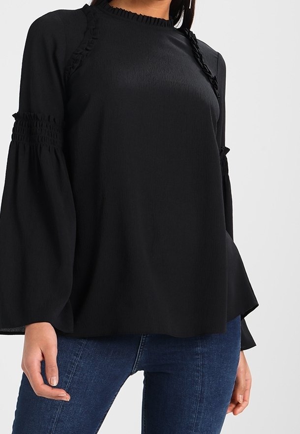 Schwarze Bluse mit Rüschenakzenten, langen Ärmeln, strukturierter Stoff und rundem Ausschnitt, kombiniert mit blauen Jeans.