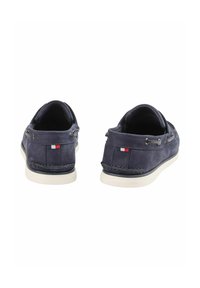 Marineblaues Wildleder-Bootsschuh-Modell mit weißen Gummisohlen, verziert mit Metallösen und einem kleinen dreifarbigen Flaggenakzent an der Seite.